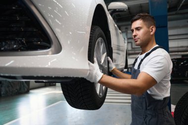 Koruyucu giysili ve eldivenli bir erkek arabanın yanında duruyor, tekerleği tutuyor, çıkarmaya çalışıyor.