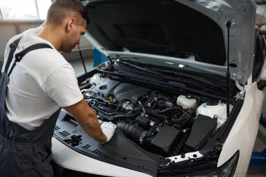 Koruyucu kıyafetli ve eldivenli tamirci arabanın yanında duruyor, tamirden önce motor bölümüne bakıyor.