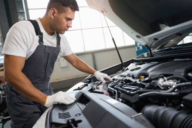 Koruyucu giysili ve eldivenli bir adam arabanın yanında duruyor, tamirden önce motor bölümüne bakıyor.