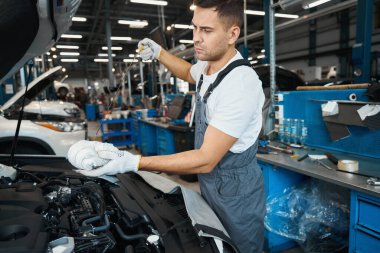 Koruyucu kıyafetli ve eldivenli tamirci kaputu açık vaziyette arabanın yanında duruyor ve motor yağı değiştiriyor.