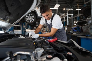 Koruyucu takım elbiseli ve eldivenli teknisyen kaputu açık arabanın yanında duruyor ve araba aküsünü kontrol ediyor.