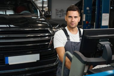 Özel takım elbiseli bir erkek, koruyucu eldivenler bilgisayarın yanında duruyor ve garajda araba tekerleklerini kontrol ediyor.