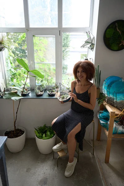Top view full length portrait of smiling female sitting on chair and holding pot plant in glass