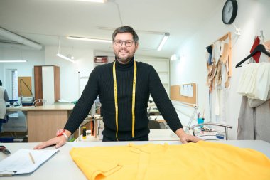 Gülümseyen terzi dikiş atölyesindeki bir kesim masasının önünde duruyor. Masadaki kumaş ve tablet.