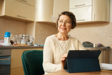 Bardaklı yaşlı kadın masada oturuyor, elinde tablet ve mutfakta alışveriş yapıyor.