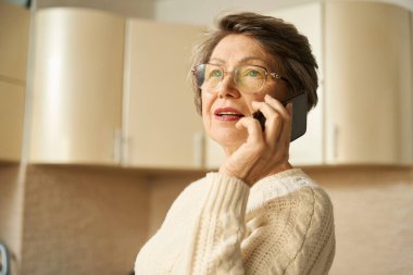 Mutfakta duran gözlüklü yaşlı kadın, elinde akıllı telefon var.