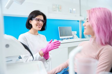 Dişçi genç bir hastayla kibarca iletişim kurar, ofis modern ekipmanlarla donatılmıştır.