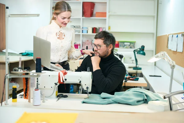 Woman manager oversees the execution of work in a sewing studio, a man looks into a laptop