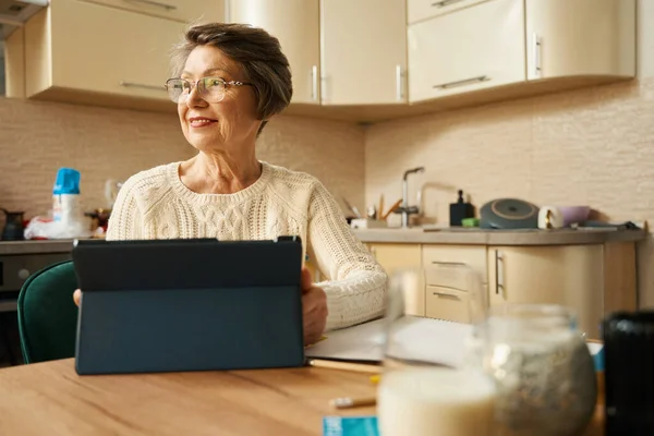 Old woman with glasses sitting at the table, holding tablet and shopping online at kitchen