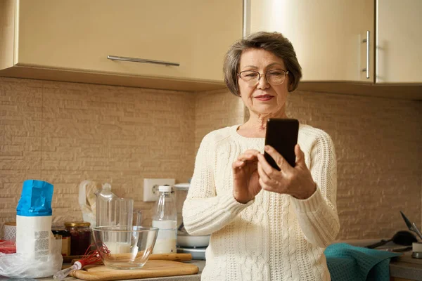 Old woman with glasses standing in the kitchen, holding smartphone and having a video call