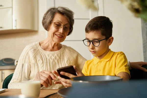 Young boy with glasses sitting at the table and holding smartphone, grandmother sitting near