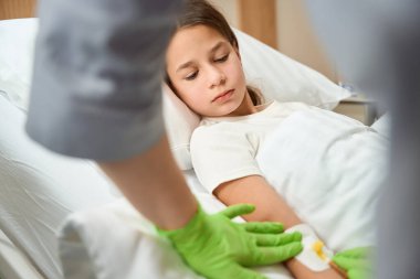 Hemşire küçük bir hastanın eline iğneyi batırıyor, kız da tıbbi manipülasyona üzülerek bakıyor.