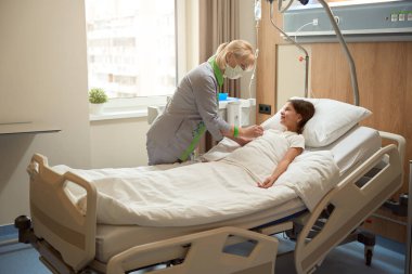 Hastane yatağında bir kızın üzerine eğilmiş koruyucu maskeli hemşire, çocuk klinikte tedavi ediliyor.