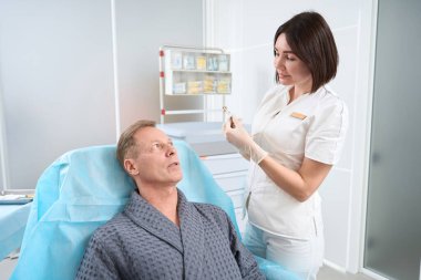 Endişeli erkek hasta elinde şırıngayla klinikte kadın bir doktorun yanında otururken endişeli görünüyor.