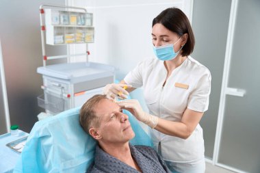 Sakin bir erkek hasta, alnına deriler arası enjeksiyon yapılırken koltuğunda sessizce oturuyor.