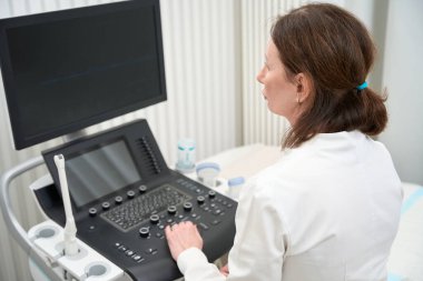Uzist doktor parlak bir ofiste ultrason makinesinin önünde oturur ve monitöre bakar.
