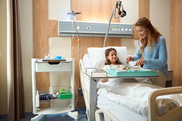 Caring mother helps a girl with a meal, a recovering girl lies on a hospital bed