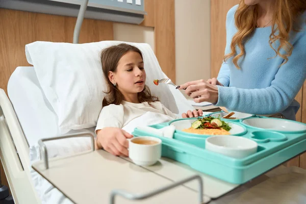 Weak girl lies on a hospital bed, mom feeds her dietary hospital food