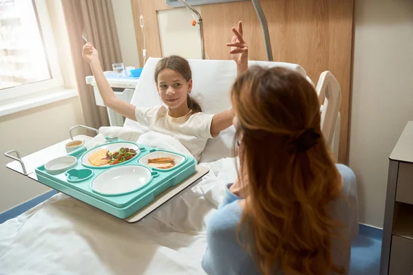 Mom and daughter in a modern hospital room, the child is having breakfast, the woman is sitting nearby