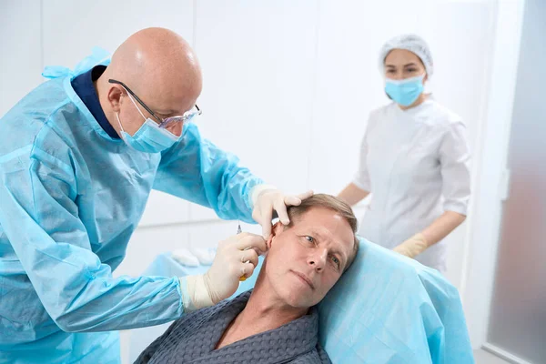 Caring doctor in surgical gown and mask desinfecting the surface on patient neck before giving an injection