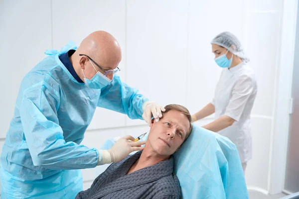 Professional doctor in full-body protective cover giving injection to male patient in armchair while nurse standing in the background