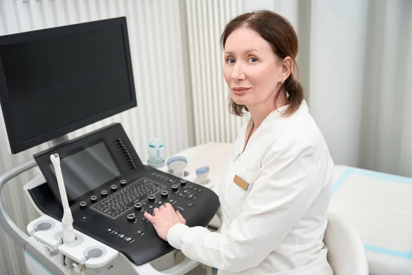 Middle-aged woman doctor sits at the workplace near the ultrasound machine and looks at the camera