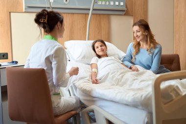 Genç hasta, annesi ve kadın doktoru hastane odasında kurnazca iletişim kuruyor, kız özel bir yatakta yatıyor.