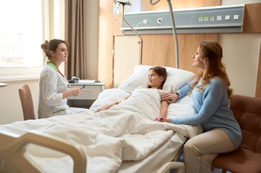 Annemle doktor, iyileşen bir kızın başucuyla iletişim kuruyor. Sağlık görevlisinin elinde kulak kanalını incelemek için otoskop var.