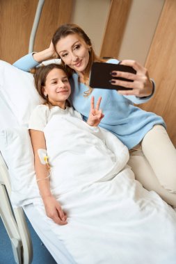 Gülümseyen anne ve kız hastane odasında muzaffer bir selfie çekiyorlar, bir kadın çocuğun yanında yatıyor.
