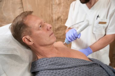 Orta yaşlı bir adam, omzunu şok dalgası terapisi için rehabilitasyon uzmanına açarken hastane yatağında yatıyor.