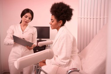 Beyaz önlüklü Afro-Amerikalı kadın jinekoloji koltuğunda oturuyor, doktor yanında duruyor ve hastaya bakıyor.