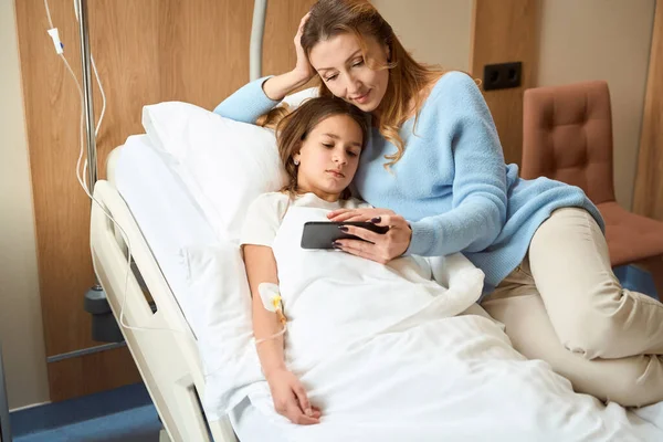 Woman and a girl lie on a special bed and look at phone screen, mother supports her daughter after surgery