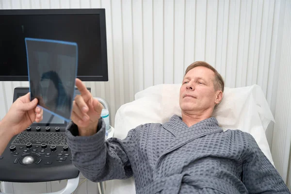 Calm middle-aged man in a hospital bed looking and pointing at a radiogram of his knee in doctor hands