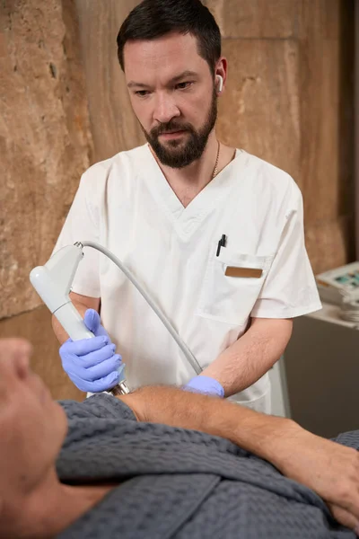 Serious brunet doctor carefully observing his patient while performing shockwave therapy on him