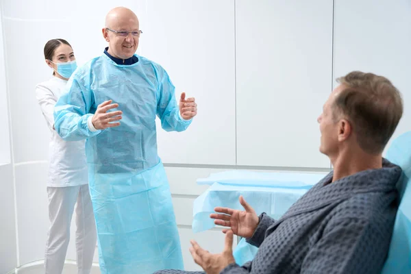 Joyful bald man in surgical suit entering the room with nurse while male patient sitting on hospital bed
