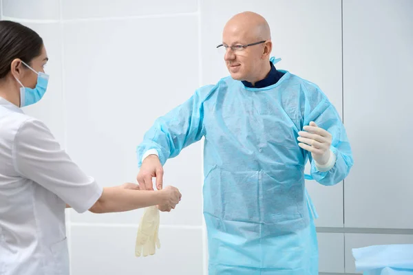 Joyous male surgeon in sterile coverall letting scrub technician put latex gloves on his hands