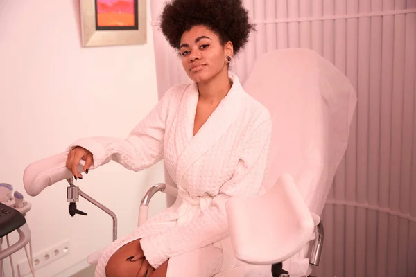 African American lady in white coat sitting on gynecological chair and looking at the camera