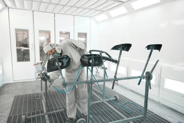 Koruyucu takım elbiseli bir adam boya kabininde duruyor, elinde sprey tabanca ve araba detaylarını boyuyor.