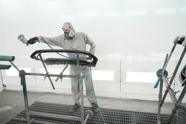 Koruyucu takım elbiseli bir erkek boya standında duruyor, elinde sprey tabanca ve boyalı araba ayrıntıları var.