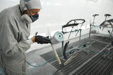 Koruyucu takım elbiseli profesyonel bir adam boya kabininde elinde sprey tabanca ve garajda araba boyası detaylarıyla dikiliyor.