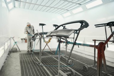 Koruyucu takım elbiseli bir adam boya kabininde duruyor, elinde sprey tabanca ve garajda araba boyası detaylarını tutuyor.