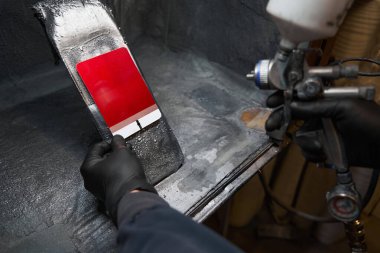 Koruyucu eldivenli bir adam boya standında duruyor, elinde sprey tabanca ve boya testi kartı tutuyor.