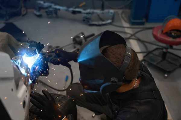 Man in a protective helmet, clothes and gloves welds metal parts of car in a workshop