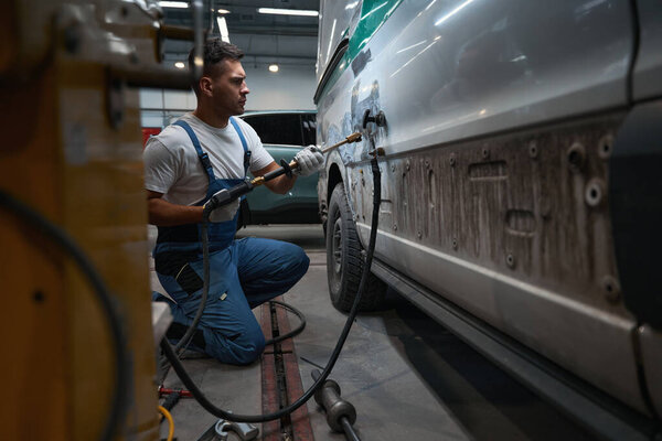 Механик в специальном костюме сидит рядом с машиной, держит специальный инструмент и снимает краску с боковой двери