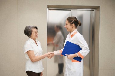 Doctor greets an elderly patient at the elevator in the lobby, they shake hands with each other