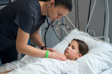 Doctor standing in ward near young patient and listening with stethoscope in the hospital