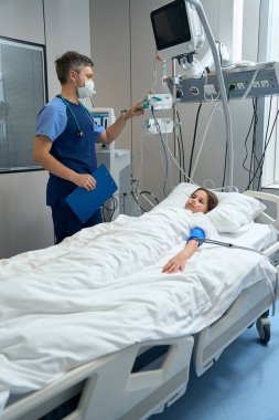 Lady lying on bed in ward, male doctor in protective mask standing near and checking information of health status