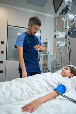 Lady lying on bed in ward, male doctor in medical clothes standing near and checking her pulse