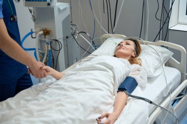 Adult female lying on bed in ward, male doctor standing near and holding her hand