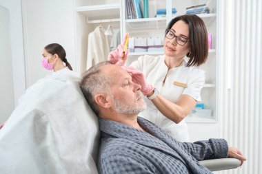 Dermatologist makes injections in the forehead of a man, he is undergoing a course of plasmolifting, an assistant is nearby
